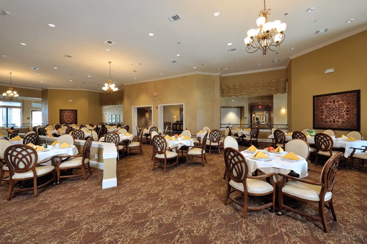 Spacious dining room with multiple round tables set with white tablecloths and yellow napkins, wooden chairs, and chandeliers.