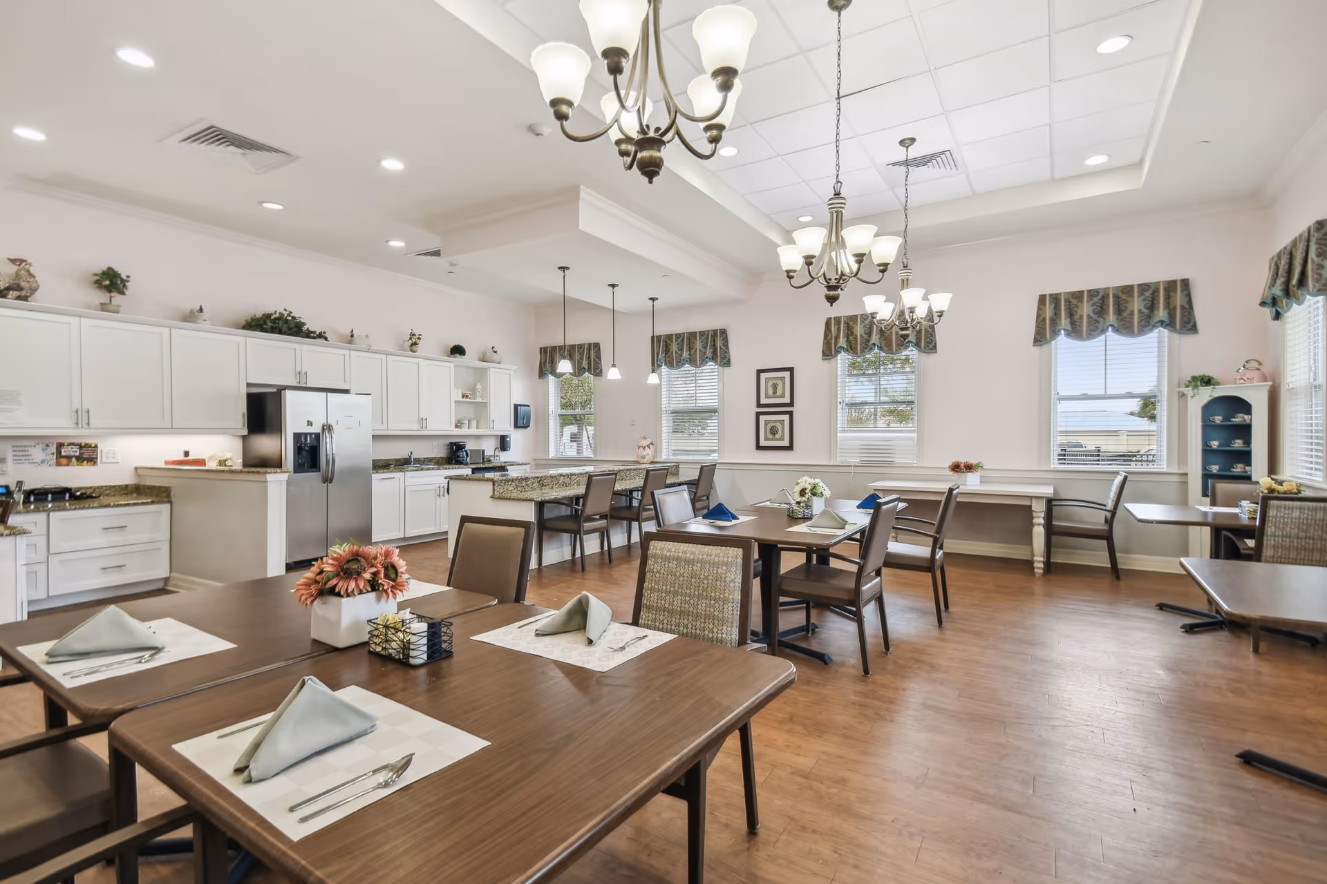 Bright communal dining room with multiple set tables and chairs, a kitchen area with white cabinets and a stainless refrigerator, and hanging chandeliers.