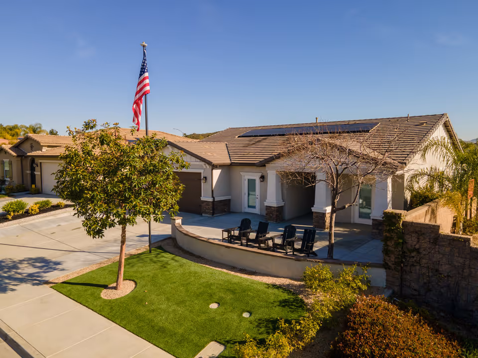 Exterior view of a single-story residential building with a tiled roof and solar panels. The front yard features a small green lawn with a tree, an American flag on a flagpole, and a patio area with four black chairs. The sky is clear and blue.