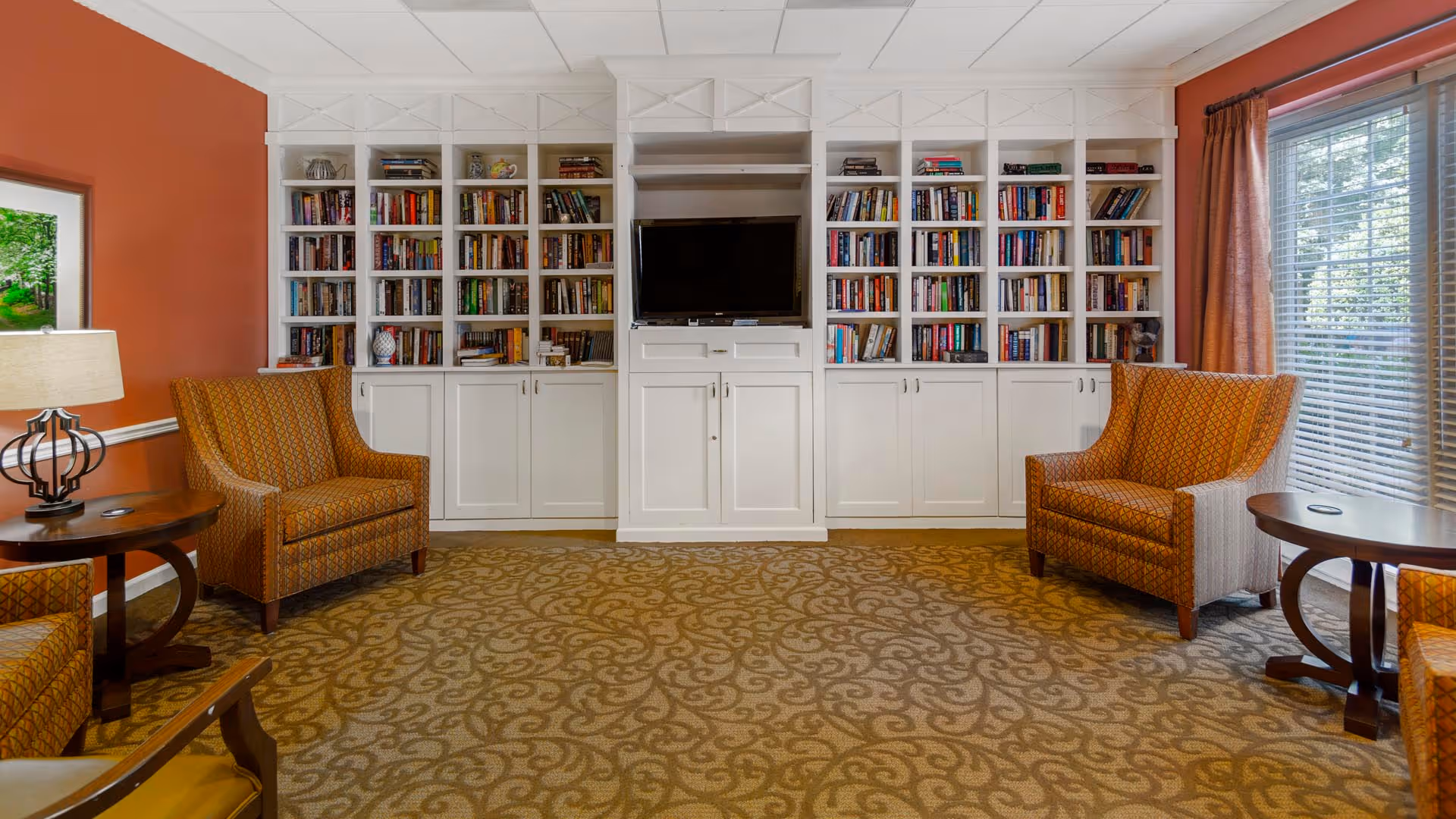 Cozy common room with built-in white bookshelves and a central TV, flanked by upholstered armchairs and side tables.
