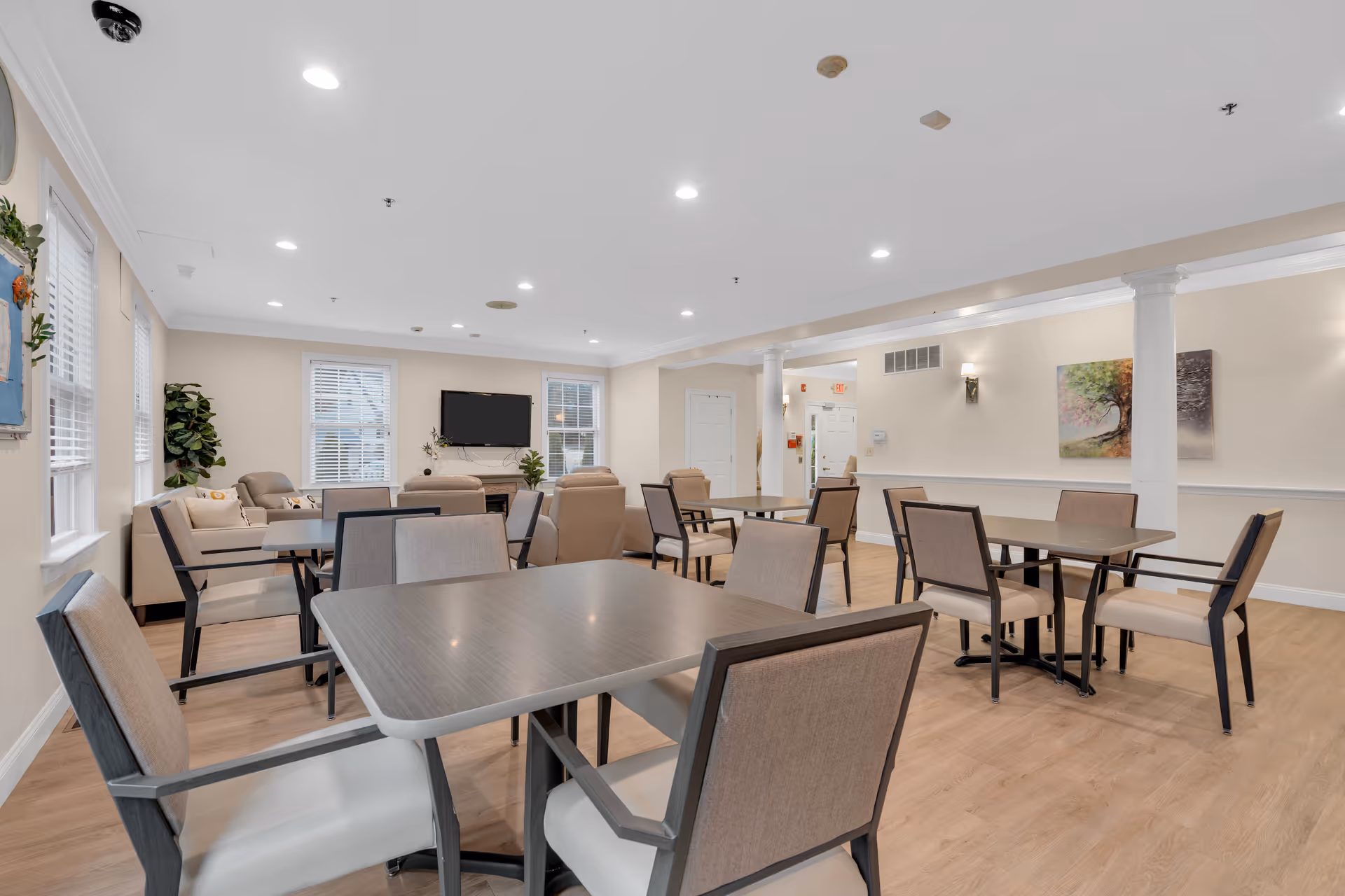 Bright communal dining/activity room with multiple tables and chairs, lounge seating, a wall-mounted TV, and light wood flooring.