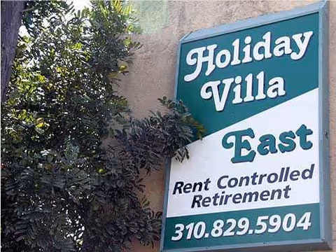 Exterior sign for Holiday Villa East Rent Controlled Retirement beside trees.