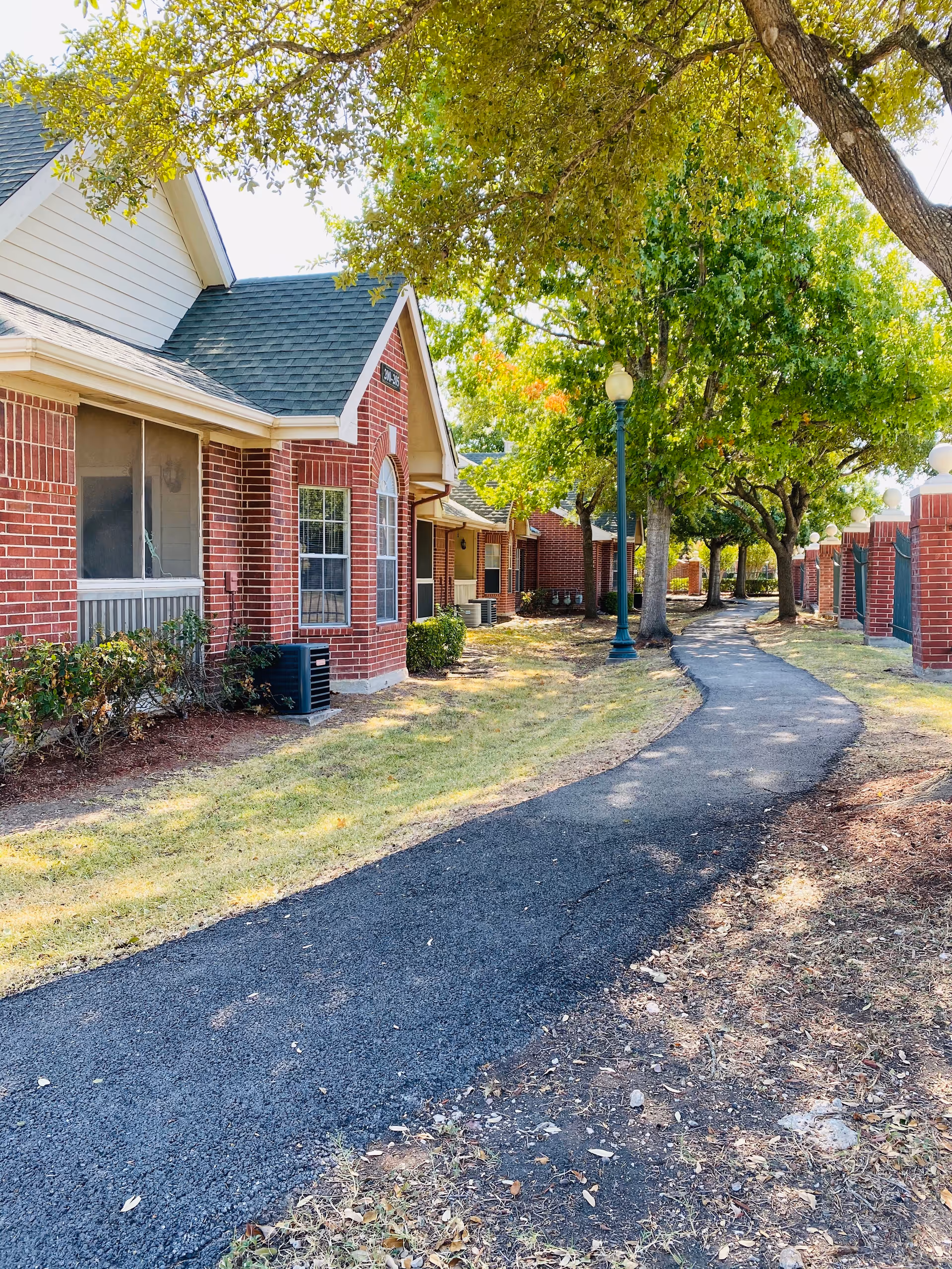 A paved walkway winding through a landscaped outdoor area with trees and grass, bordered by red brick buildings with green roofs and windows. There are lamp posts along the path providing lighting.