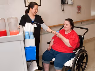 A healthcare worker in black scrubs is smiling and holding the hand of a woman in a red shirt sitting in a wheelchair in a hallway. There is a cart with pitchers and cups nearby.