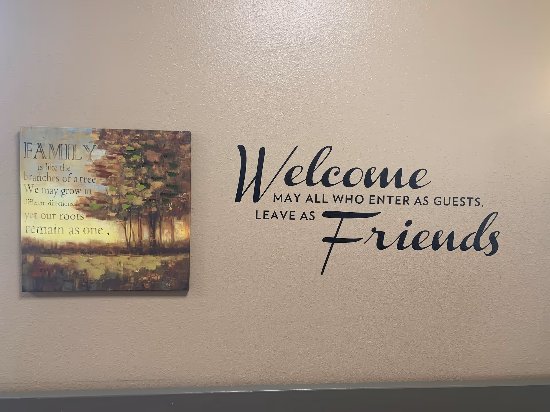 A beige wall with two pieces of wall decor. On the left is a framed painting of trees with the text: 'FAMILY is like the branches of a tree We may grow in different directions yet our roots remain as one.' On the right is a large wall decal with the words: 'Welcome MAY ALL WHO ENTER AS GUESTS, LEAVE AS Friends'.