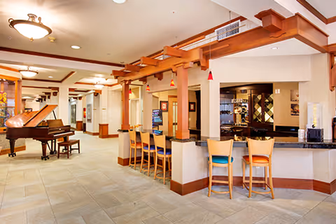 Interior view of a senior living facility common area featuring a grand piano on the left, a bar-style counter with wooden chairs in the center, and warm lighting fixtures on the ceiling. The space has tiled flooring and wooden architectural accents.