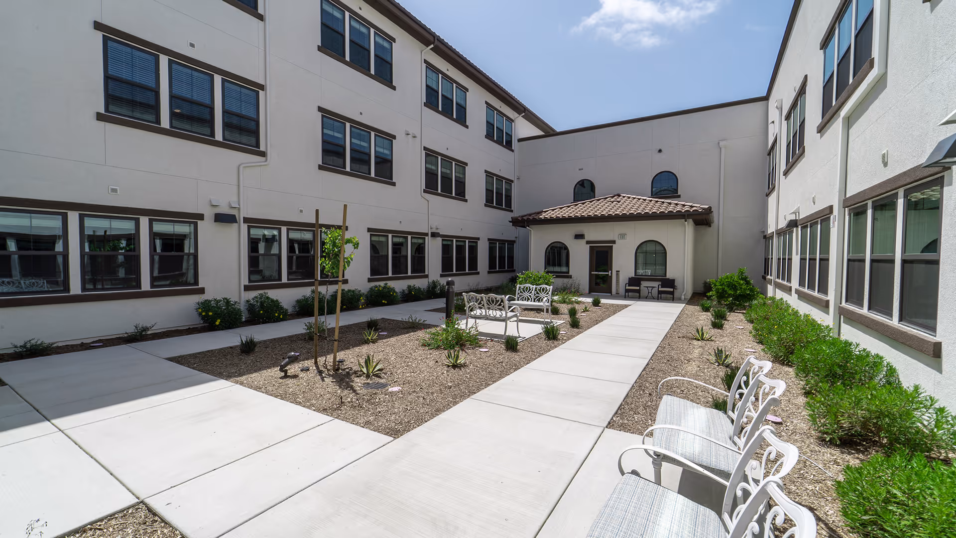 Outdoor courtyard area at La Marea Senior Living with white benches, small plants, and a paved walkway surrounded by a three-story building with multiple windows under a partly cloudy sky.