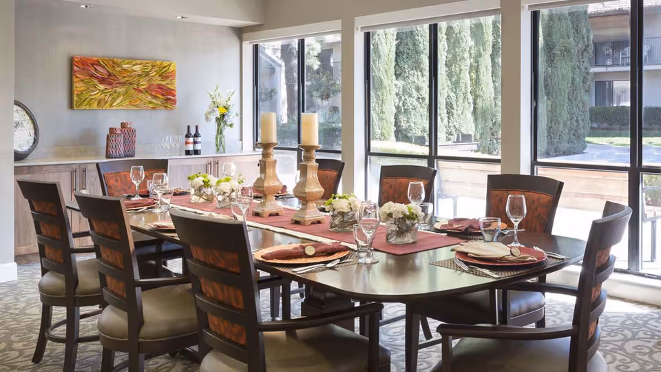 A well-lit dining room with a large rectangular wooden table set for eight people. The table is decorated with a red runner, floral centerpieces, two large candles, and place settings including plates, napkins, and glassware. Large windows provide a view of tall green trees outside. There is a sideboard against the wall with decorative items and a colorful abstract painting above it.