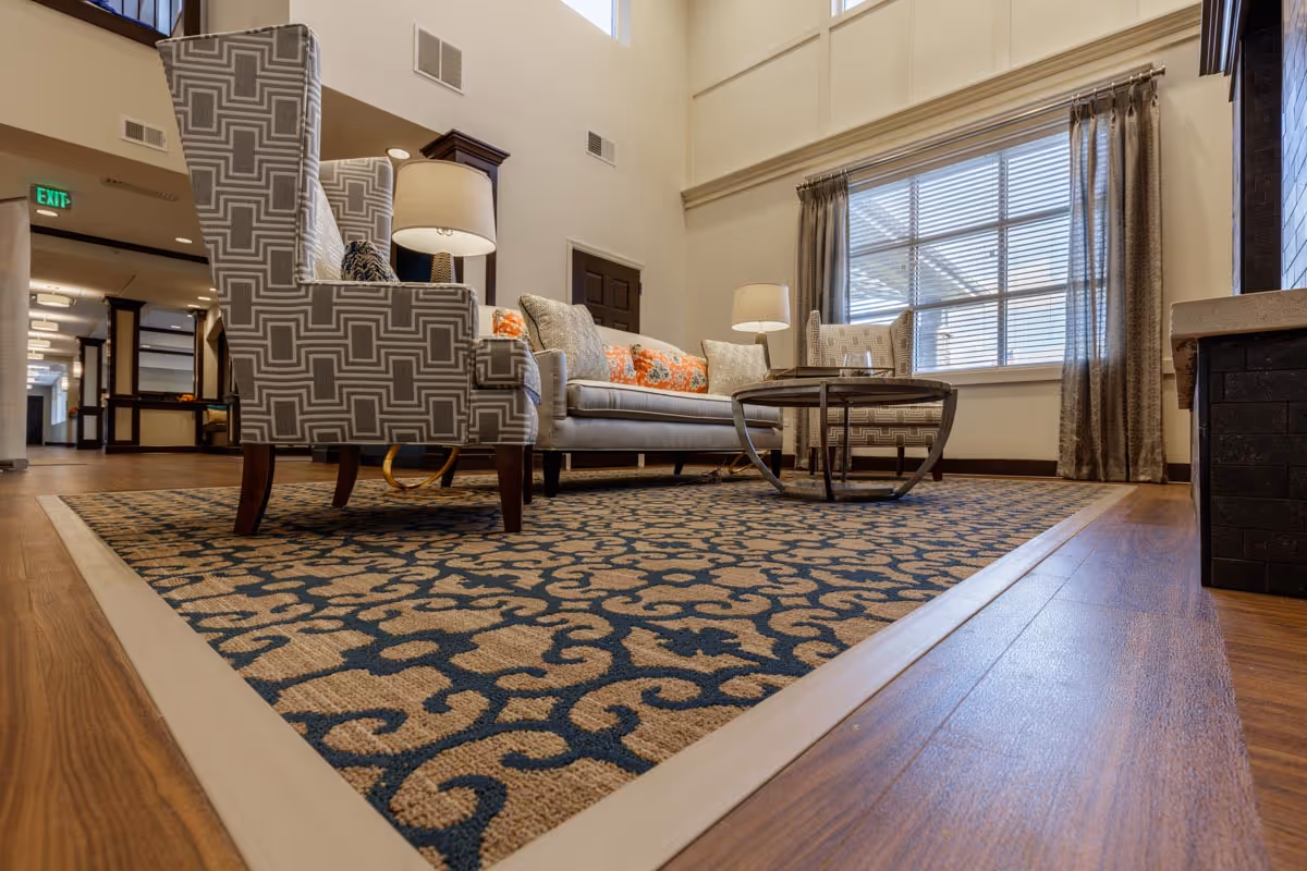 A cozy living room area with a patterned area rug, two high-back armchairs with geometric designs, a sofa with decorative pillows, a round coffee table, two table lamps, large window with curtains, and wooden flooring. The room has high ceilings and neutral-colored walls.