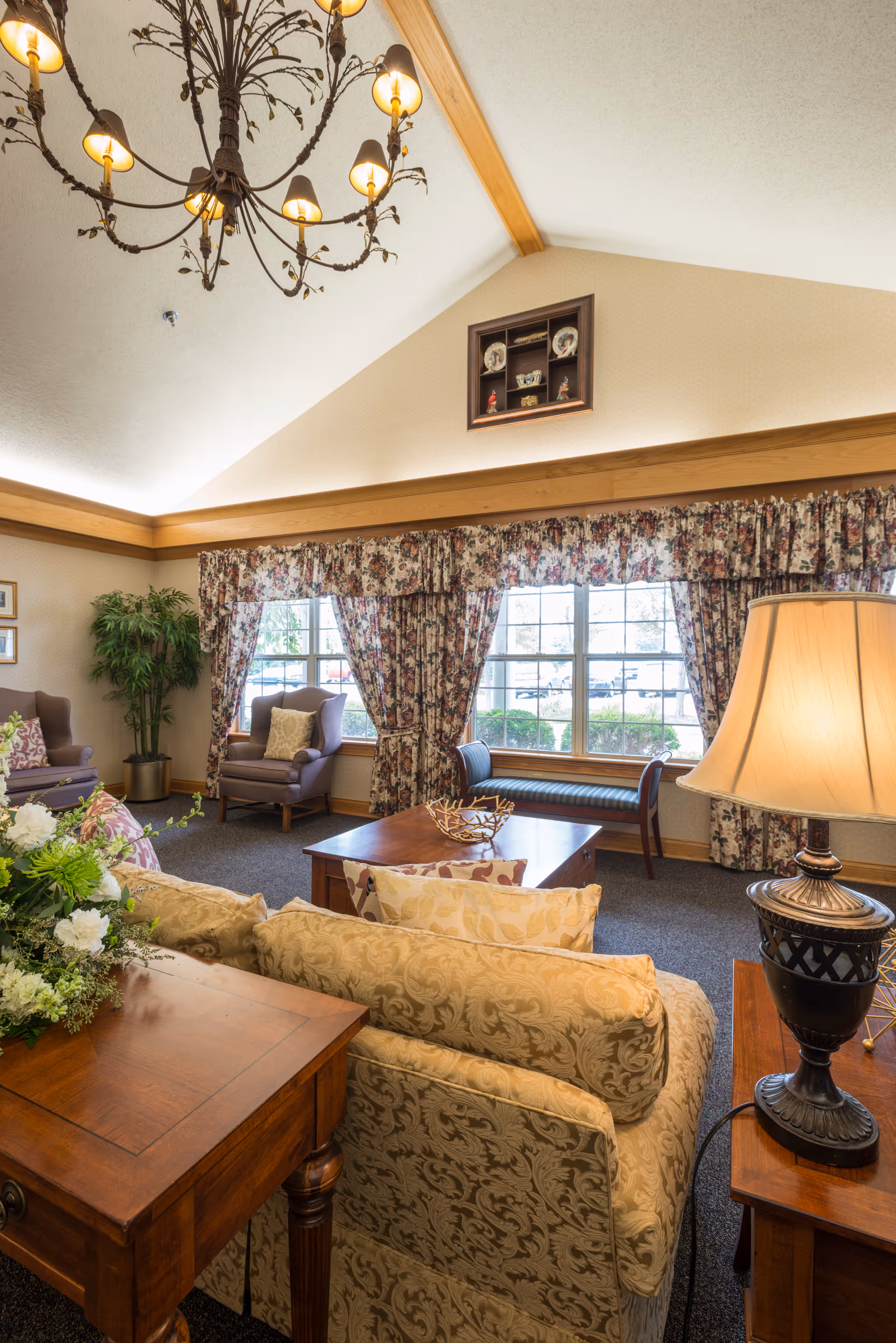 Sunlit lounge area with upholstered sofas and chairs, a coffee table, floral curtains, and a chandelier.