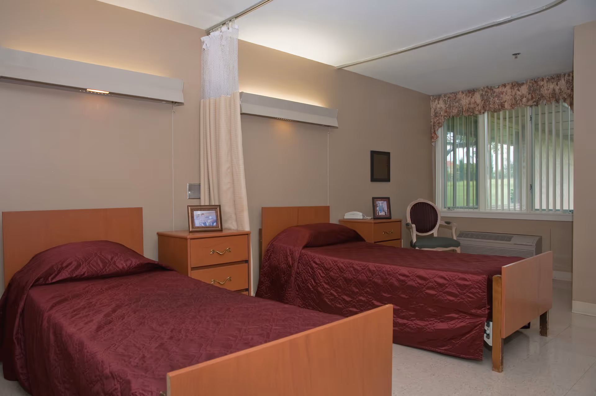A room with two single beds covered in maroon bedspreads, separated by a wooden nightstand with framed photos on top. There is a curtain divider between the beds, a window with vertical blinds and floral valance, a chair, and an air conditioning unit below the window.