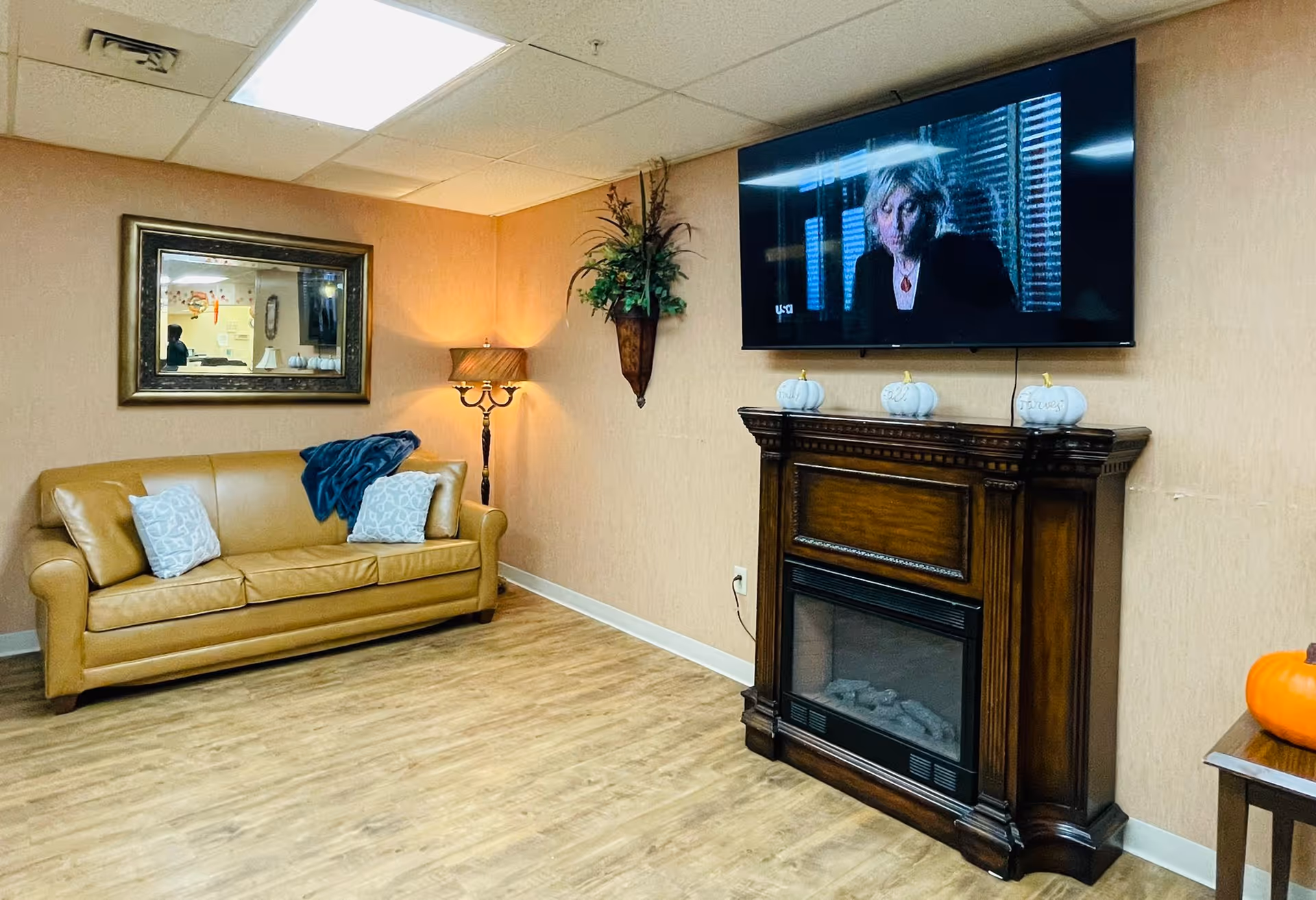 Cozy common room with a tan leather sofa, decorative pillows and lamp, a wall mirror, and a mounted TV above a wooden electric fireplace.