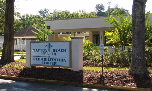 Exterior view of Daytona Beach Health and Rehabilitation Center showing the facility's sign in front of a landscaped area with trees and bushes, and part of the building entrance in the background.