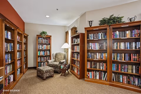 A cozy reading nook in a senior living facility featuring wooden bookshelves filled with books, a patterned armchair with a matching ottoman, a floor lamp, and a small wooden magazine rack. The walls are painted in neutral tones with one accent wall in a warm reddish color.