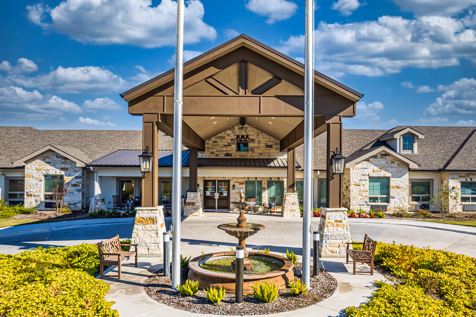 Entrance of a senior living facility with a covered porte-cochere, central fountain, benches, and manicured landscaping under a blue sky.