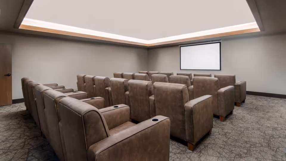 Small screening room with rows of brown upholstered recliner chairs facing a blank projection screen under recessed ceiling lighting.