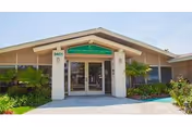 Exterior front entrance of Long Beach Care Center, a single-story building with a peaked roof, glass double doors, and a green sign above the entrance. The building is surrounded by plants and shrubs.
