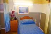 A small nursing center bedroom with a single bed covered in a blue quilt. Next to the bed is a wooden nightstand with a telephone and a vase with red flowers. A framed picture hangs on the wall above the bed. The room has light-colored walls with a two-tone paint design and a curtain partially visible on the left side.