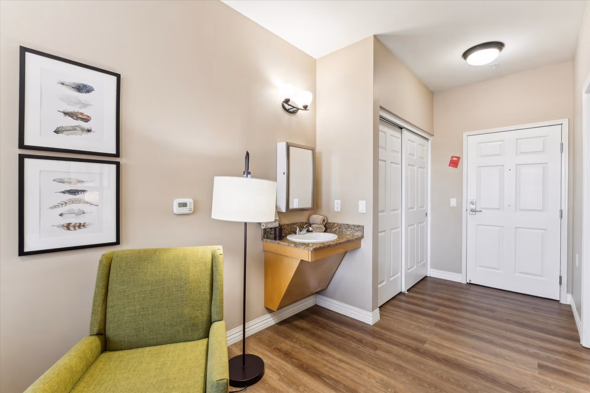 Interior view of a room corner featuring a green armchair, a floor lamp, two framed feather artworks on the wall, a small wall-mounted sink with a mirror above it, and a white door with adjacent closet doors. The floor is wood and the walls are painted beige.