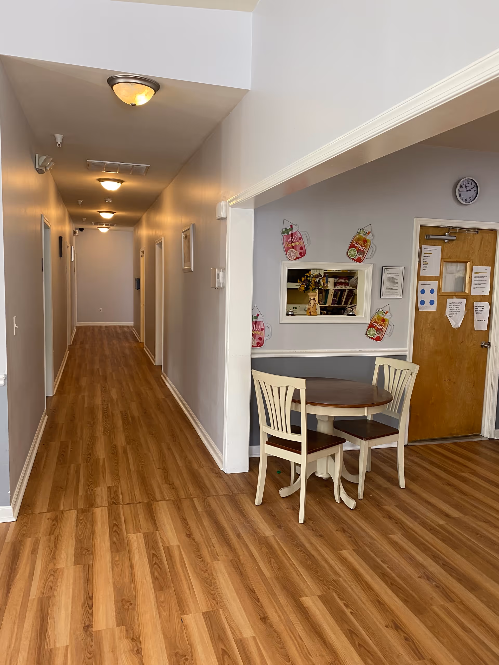 A hallway with wooden flooring and several closed doors on the left side. On the right side, there is a small seating area with a round wooden table and two white chairs. The wall behind the seating area has a small window and decorative signs with colorful drink illustrations. A wooden door with papers attached and a clock above it is visible in the background.