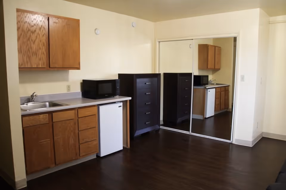 Small studio kitchenette with wood cabinets, a sink, mini fridge and microwave next to a dark chest of drawers and sliding mirrored closet doors.