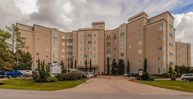 Front exterior of Pegasus Landing of Tanglewood, a beige multi-story senior living building with parking.
