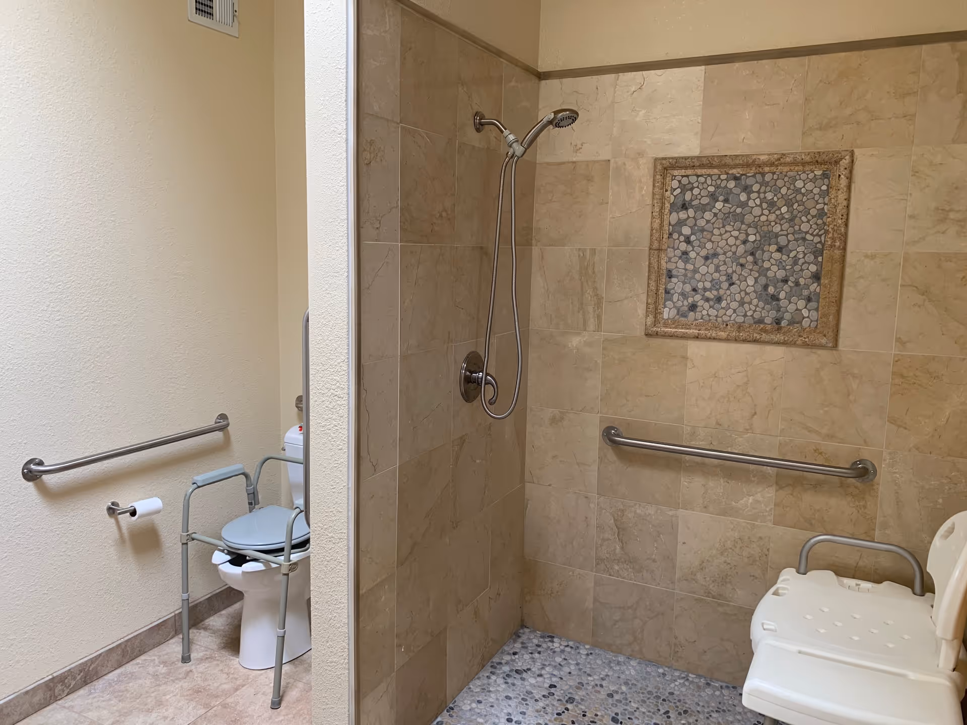 A bathroom with beige tiled walls and floor. There is a walk-in shower with a handheld showerhead and a pebble stone tile accent on the wall. A white shower chair is placed inside the shower. To the left, there is a toilet with a support frame around it and a grab bar on the wall next to it. A toilet paper holder is mounted on the wall near the toilet.