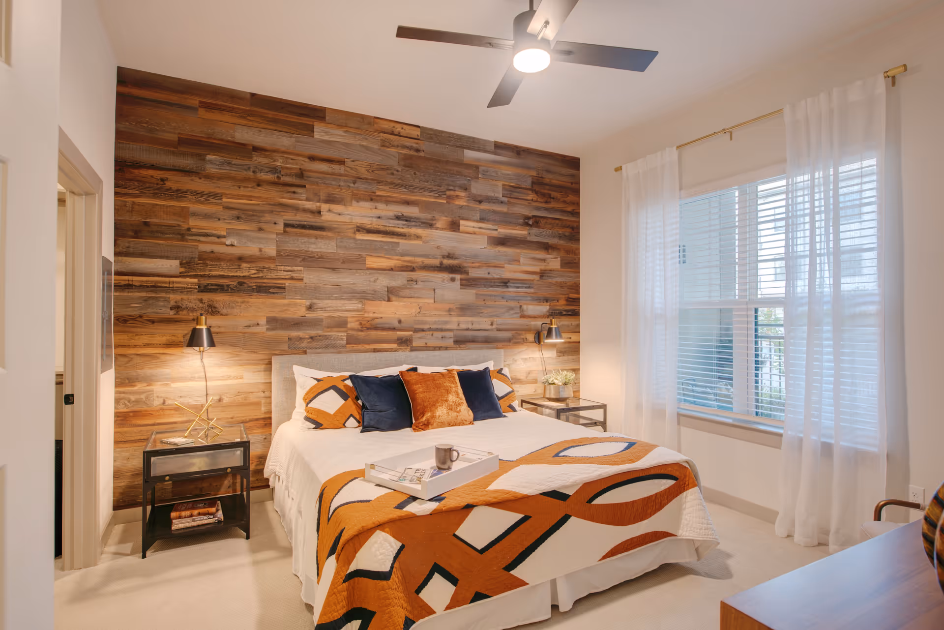 A cozy bedroom with a large bed featuring a white and orange geometric patterned quilt and multiple pillows. The back wall is covered with rustic wooden panels. There are two black bedside tables with lamps on each side of the bed. A ceiling fan with a light is mounted on the ceiling. A large window with white sheer curtains allows natural light into the room.