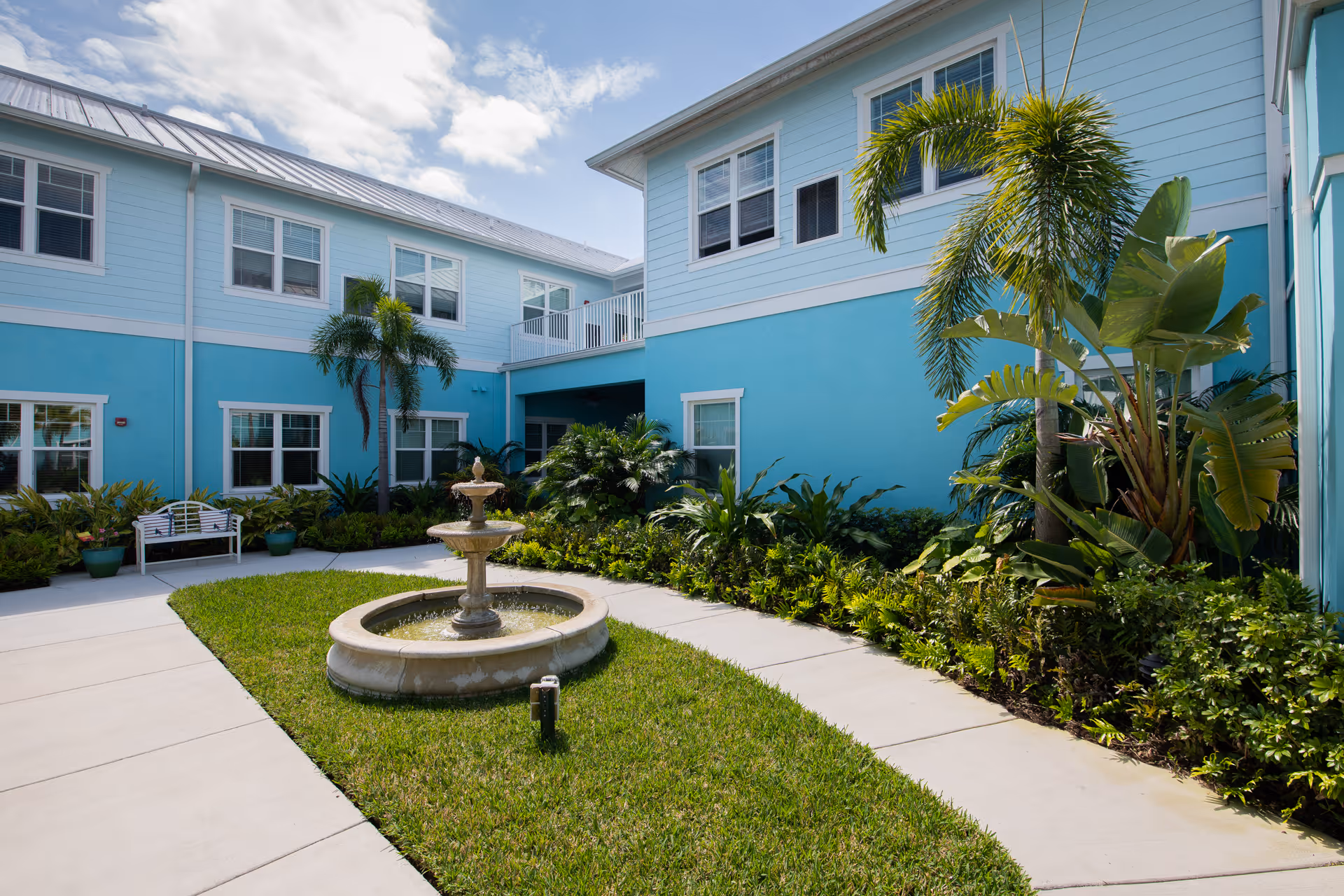 Sunny courtyard with a central fountain, tropical plants, a bench, and blue two-story building facades.