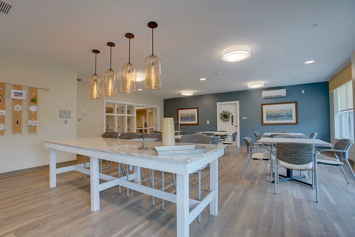 A bright and modern dining room with several tables and chairs arranged for seating. A large white table with a marble top is in the foreground, with four pendant lights hanging above it. The walls are painted in neutral tones with one accent wall in blue, decorated with framed artwork and a wreath on a door. The room has large windows allowing natural light to enter.