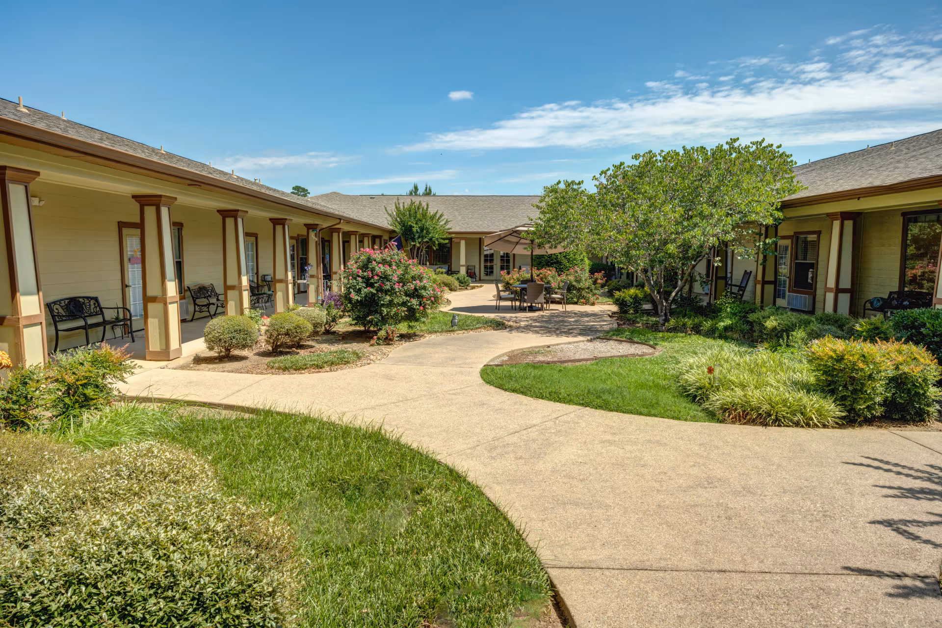 Sunny landscaped courtyard with paved walkways, seating areas, shrubs and a single-story assisted living building surrounding it.