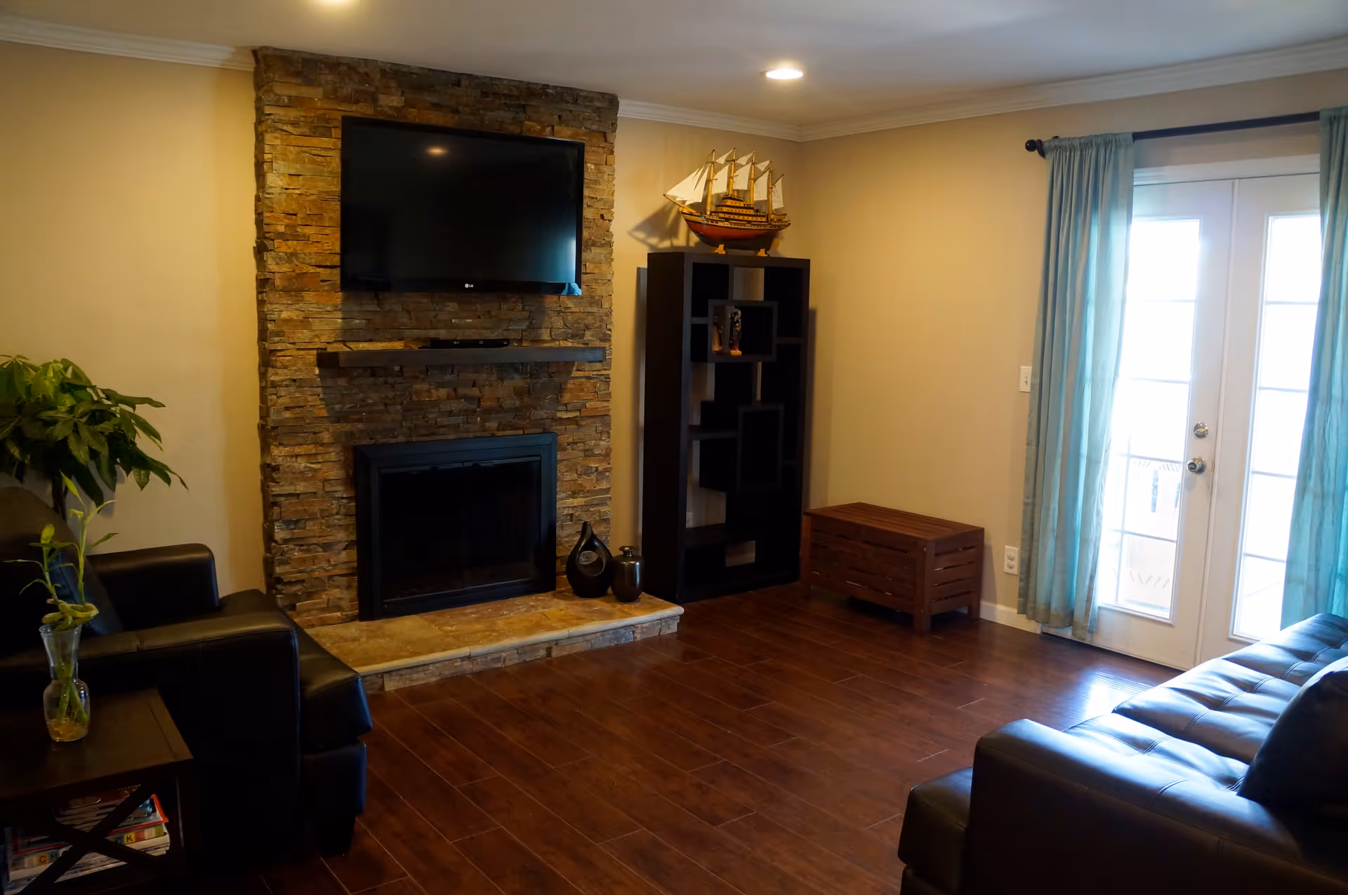 Cozy living room with a stone fireplace and wall-mounted TV, dark leather sofas, shelving unit, and French doors with blue curtains.