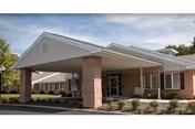 Covered porte-cochère entrance of a brick care facility with columns, windows, and low landscaping beds.