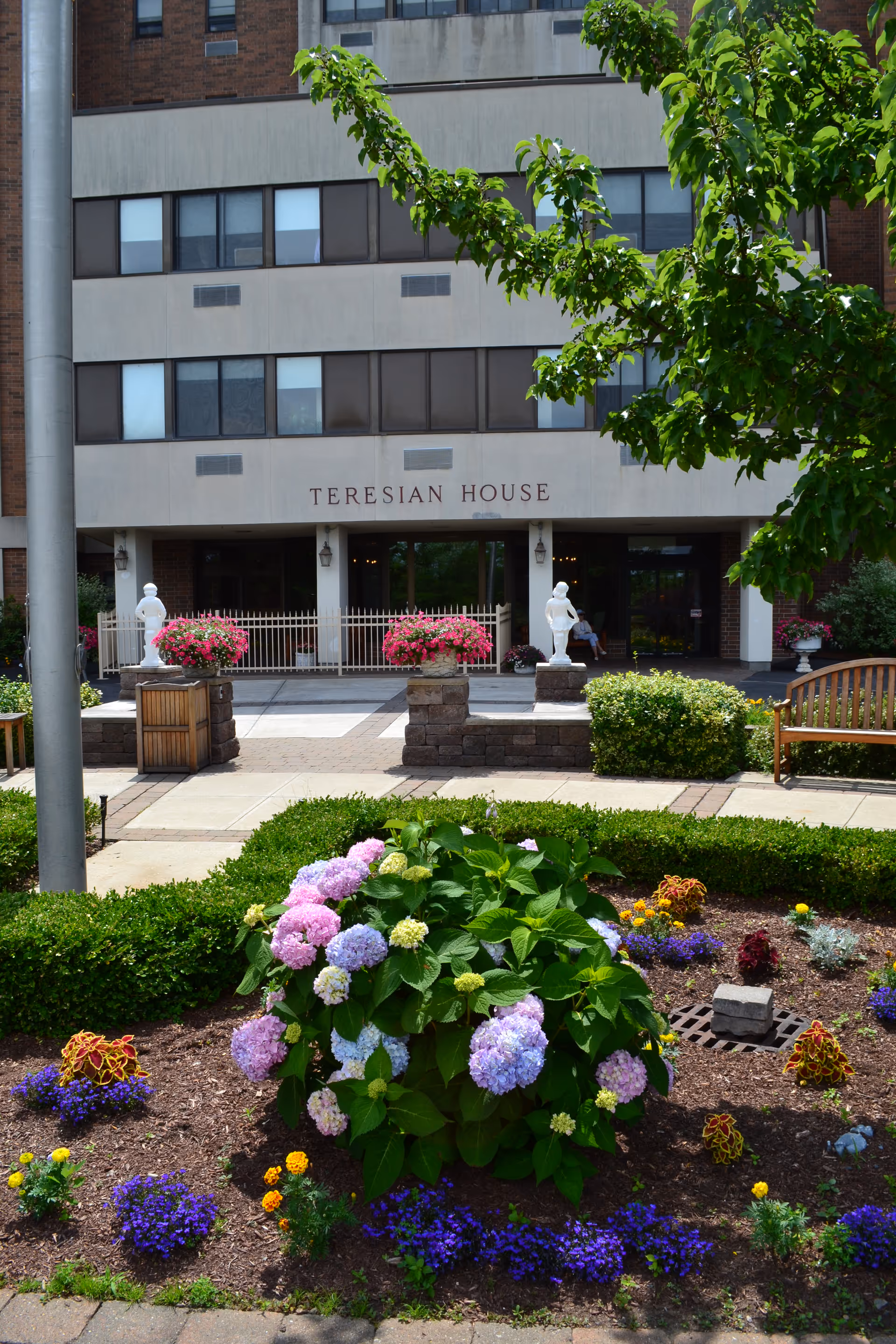 Front entrance of Teresian House with landscaped flowerbeds, benches, and the building's name above the doorway.