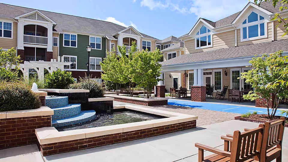 Outdoor courtyard area at a senior living facility with a small water fountain, brick planters with trees and shrubs, benches, and a covered patio with rocking chairs and tables. The building has multiple stories with beige and green siding and white trim under a blue sky with some clouds.