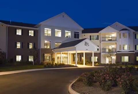 Exterior view of a multi-story senior living facility named Willow Brook at Delaware Run at dusk, with lights glowing from the windows and a covered entrance with columns. There are landscaped bushes and flowers in front of the building along a curved driveway.