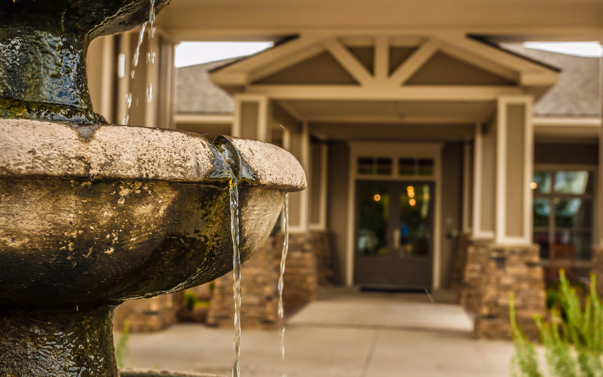 Close-up view of a stone water fountain with water flowing from its upper tier, set in front of the entrance to a building with stone pillars and a covered porch.