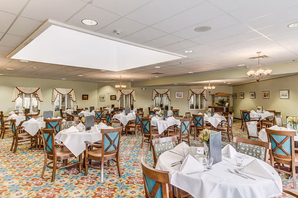 Spacious dining room with multiple round tables set with white tablecloths, wooden chairs, chandeliers, and a colorful patterned carpet.