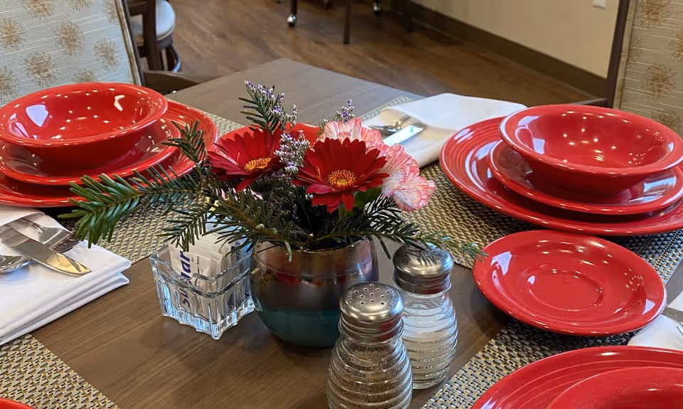 A dining table set with red plates, bowls, and saucers on woven placemats. There is a small floral centerpiece with red and pink flowers and green foliage in a vase, along with salt and pepper shakers and a glass container holding sugar packets. White napkins with silverware are placed beside the plates.
