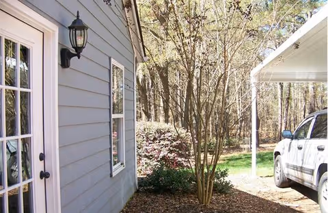 Side view of a building with gray siding, a white-framed glass door, a window, and an outdoor wall lantern. There is a tree and bushes in a mulched area next to the building, and a white vehicle is partially visible parked under a carport. The background shows a wooded area with tall trees.