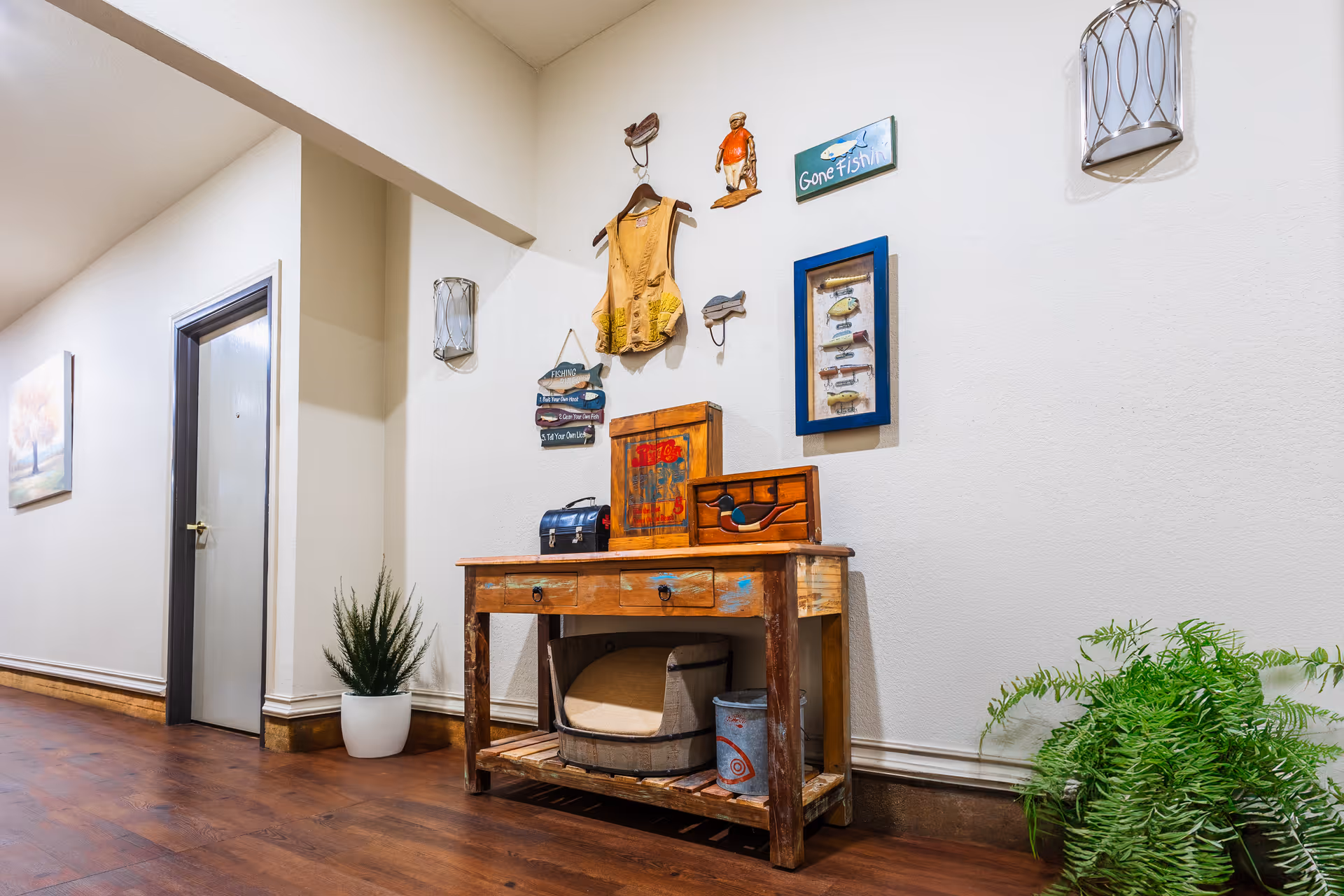 A hallway with wooden flooring and white walls featuring a rustic wooden table with drawers. On the table are decorative items including a small chest and a wooden duck. Above the table, fishing-themed wall decorations are displayed, including a vest, a small figure, and signs. There are two wall sconces on either side of the decorations. A potted plant is placed on the floor to the right of the table, and another plant is near a closed door on the left side of the hallway.