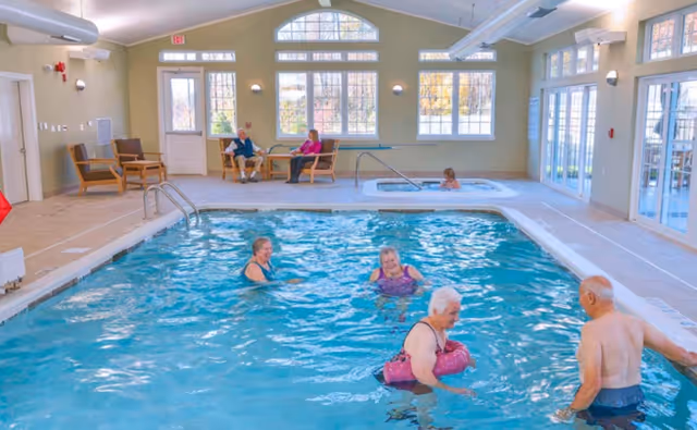 Indoor swimming pool area with four elderly people enjoying the water. In the background, two elderly people are seated and conversing near large windows that let in natural light. The room has light-colored walls, multiple windows, and a door leading outside.