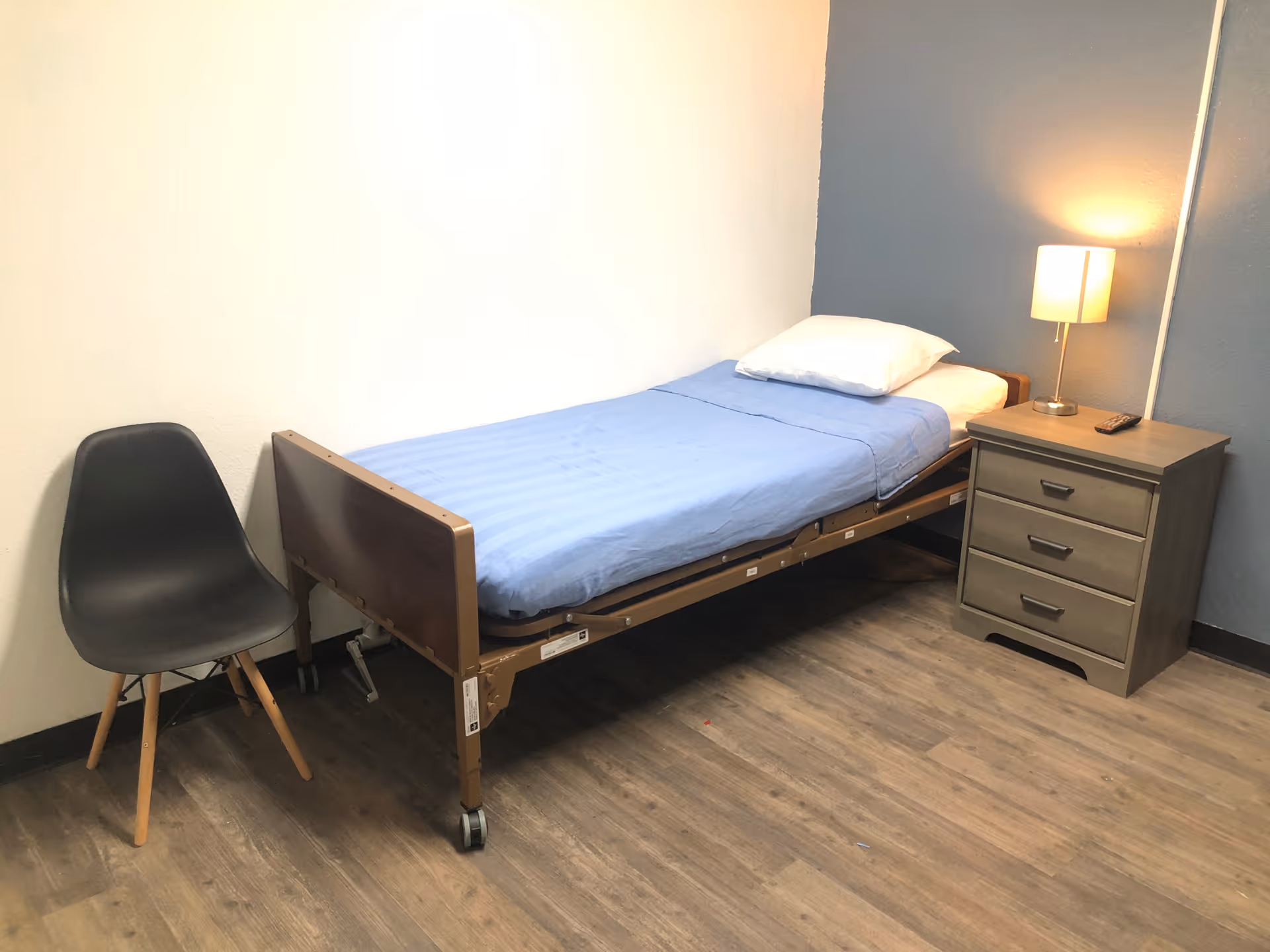 A simple room with a single hospital-style bed with a blue sheet and white pillow, a small wooden nightstand with three drawers and a lit lamp on top, and a black chair with wooden legs. The floor is wood laminate, and the walls are painted white and gray.
