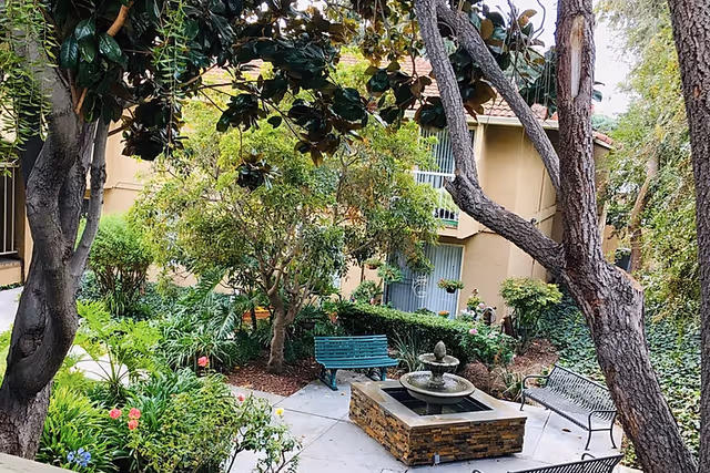A peaceful outdoor courtyard area at Villa Serena Retirement Community featuring a stone water fountain in the center, surrounded by benches and lush green trees and plants. The background shows part of the building with windows and balconies.
