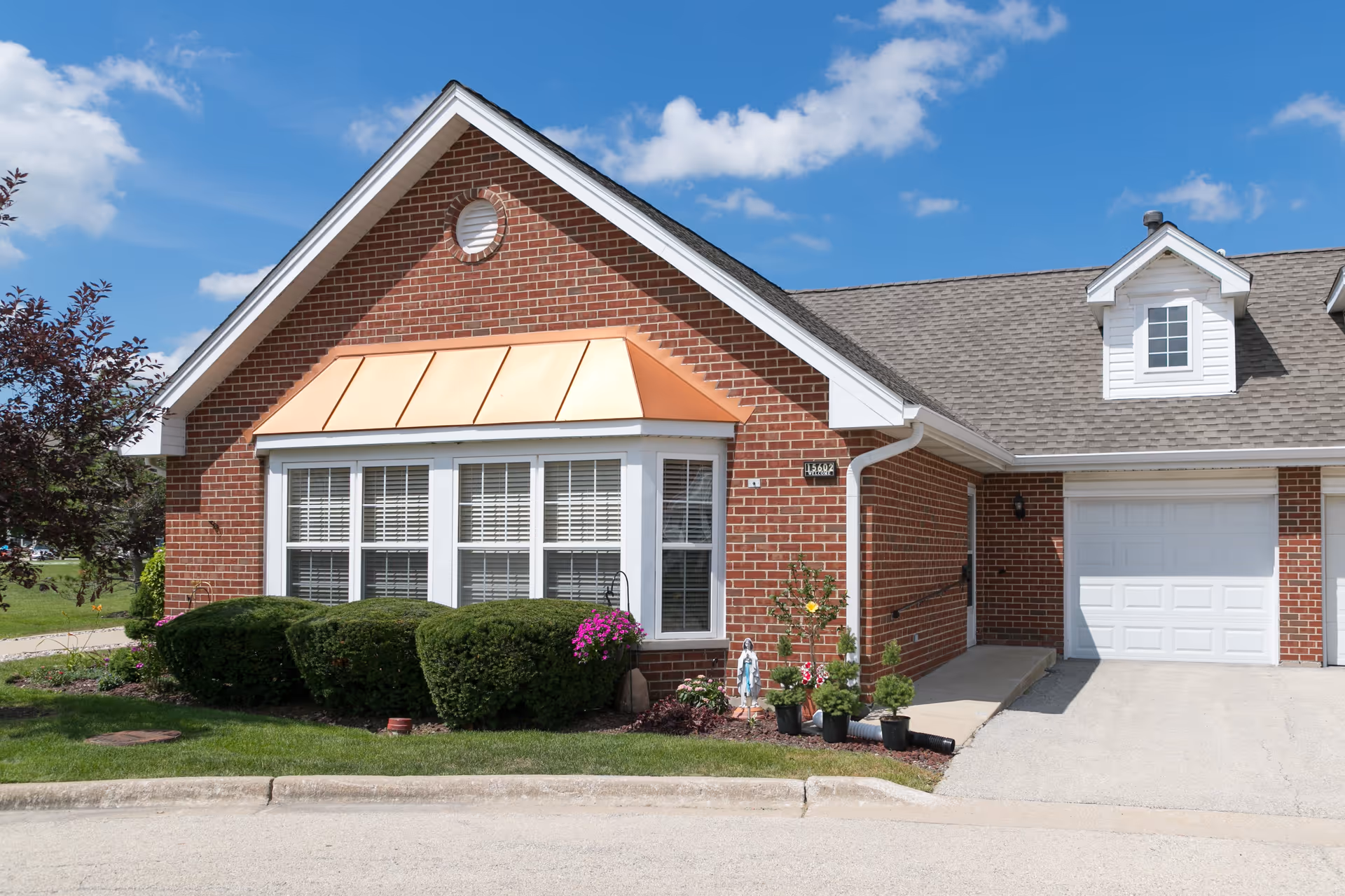 Exterior view of a brick building with a peaked roof, white-framed windows with blinds, a copper-colored awning, and a garage door. There are neatly trimmed bushes and potted plants near the building, under a blue sky with some clouds.