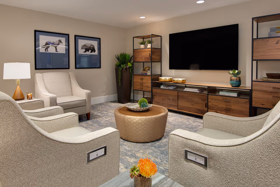 Cozy living room with armchairs arranged around a round ottoman, a wall-mounted TV, and wooden media cabinets.