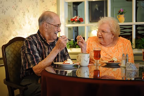 Two elderly people sit at a dining table smiling and sharing dessert.