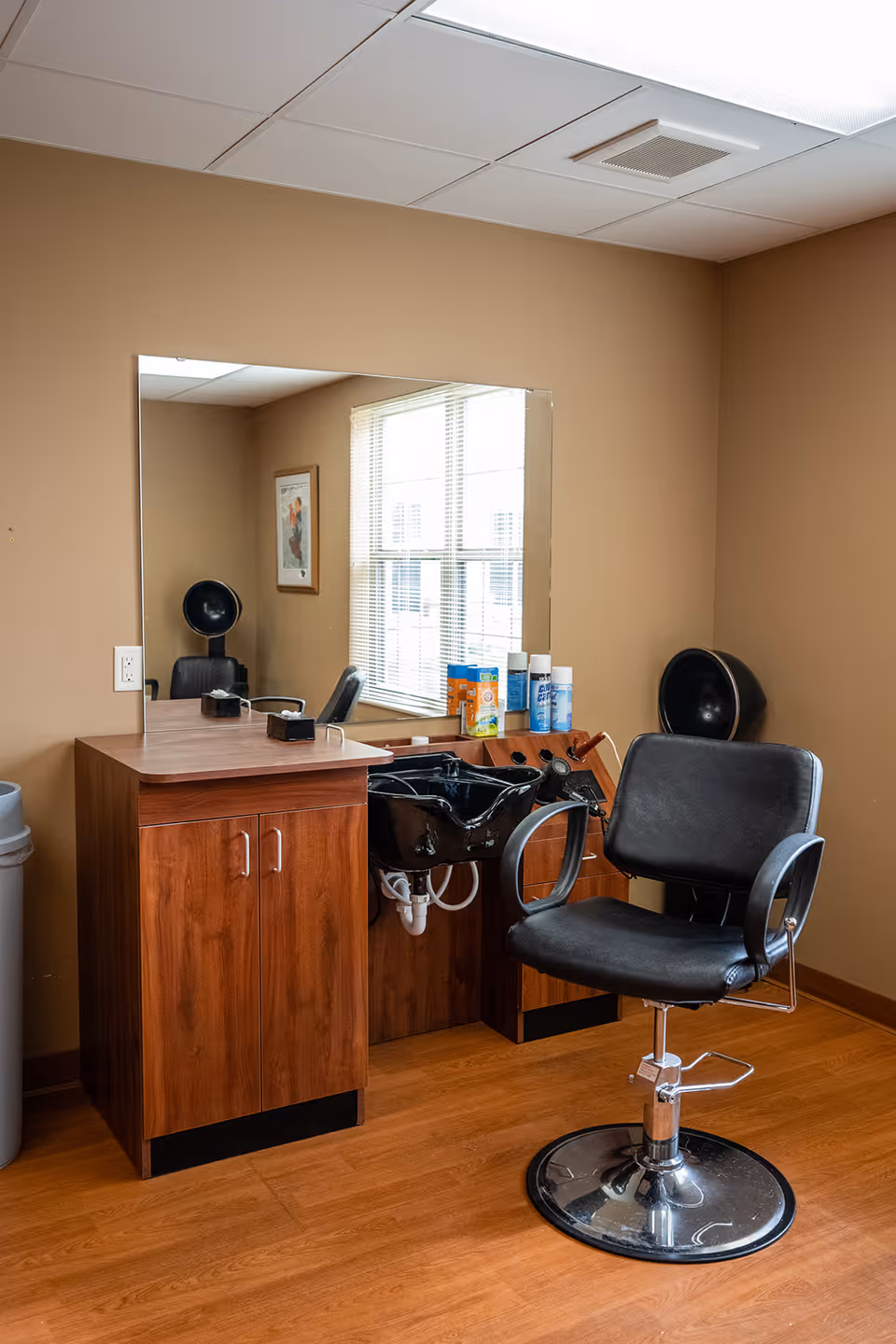 Interior view of a small hair salon area with a black salon chair, a wooden cabinet with a sink for washing hair, a large mirror on the wall, and hair care products on the counter. The room has beige walls and a window with blinds letting in natural light.