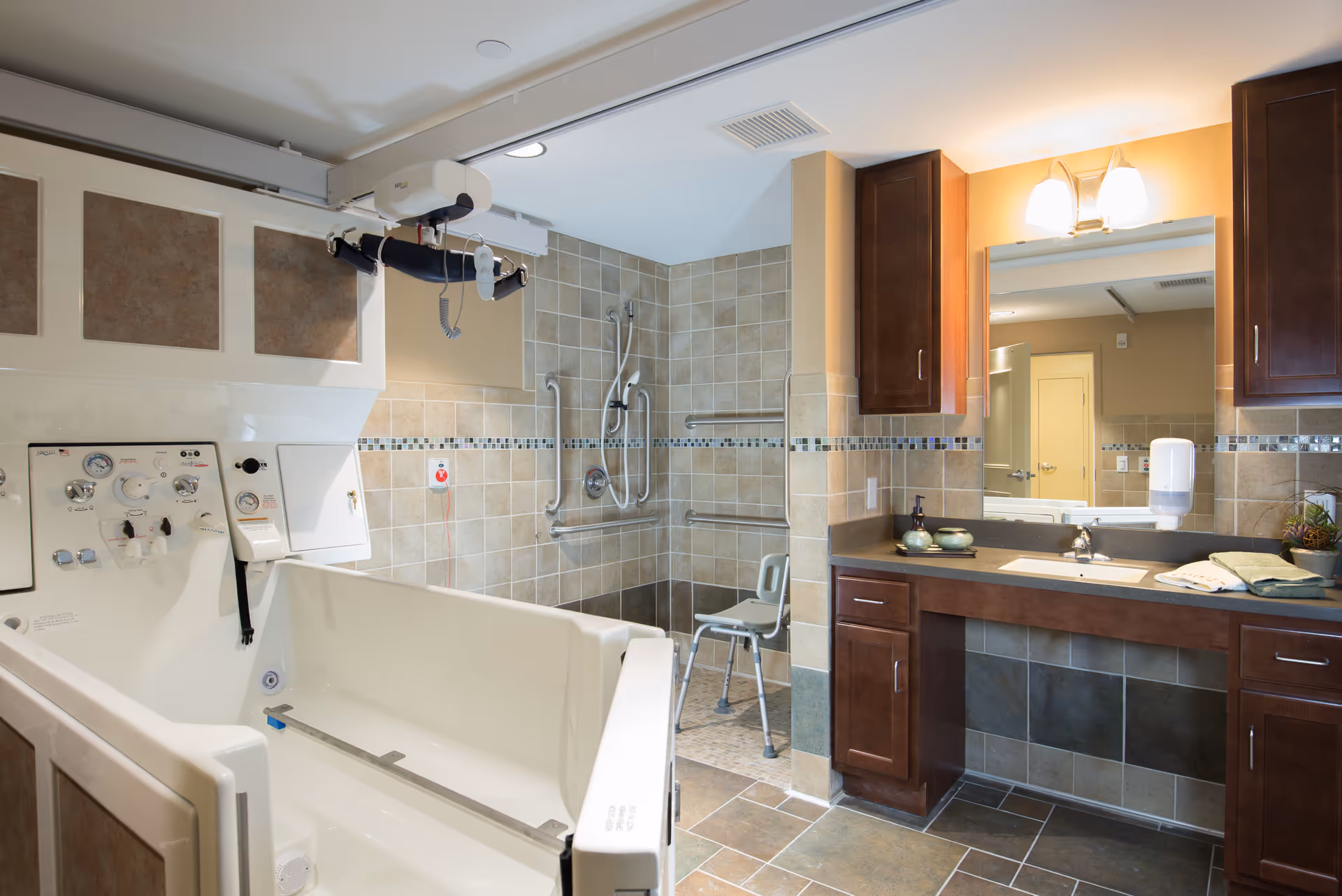 A spacious, accessible bathroom featuring a large walk-in bathtub with control panel, a tiled walk-in shower with grab bars and a shower chair, and a vanity with a sink, mirror, and wooden cabinets. The room is well-lit with warm lighting.