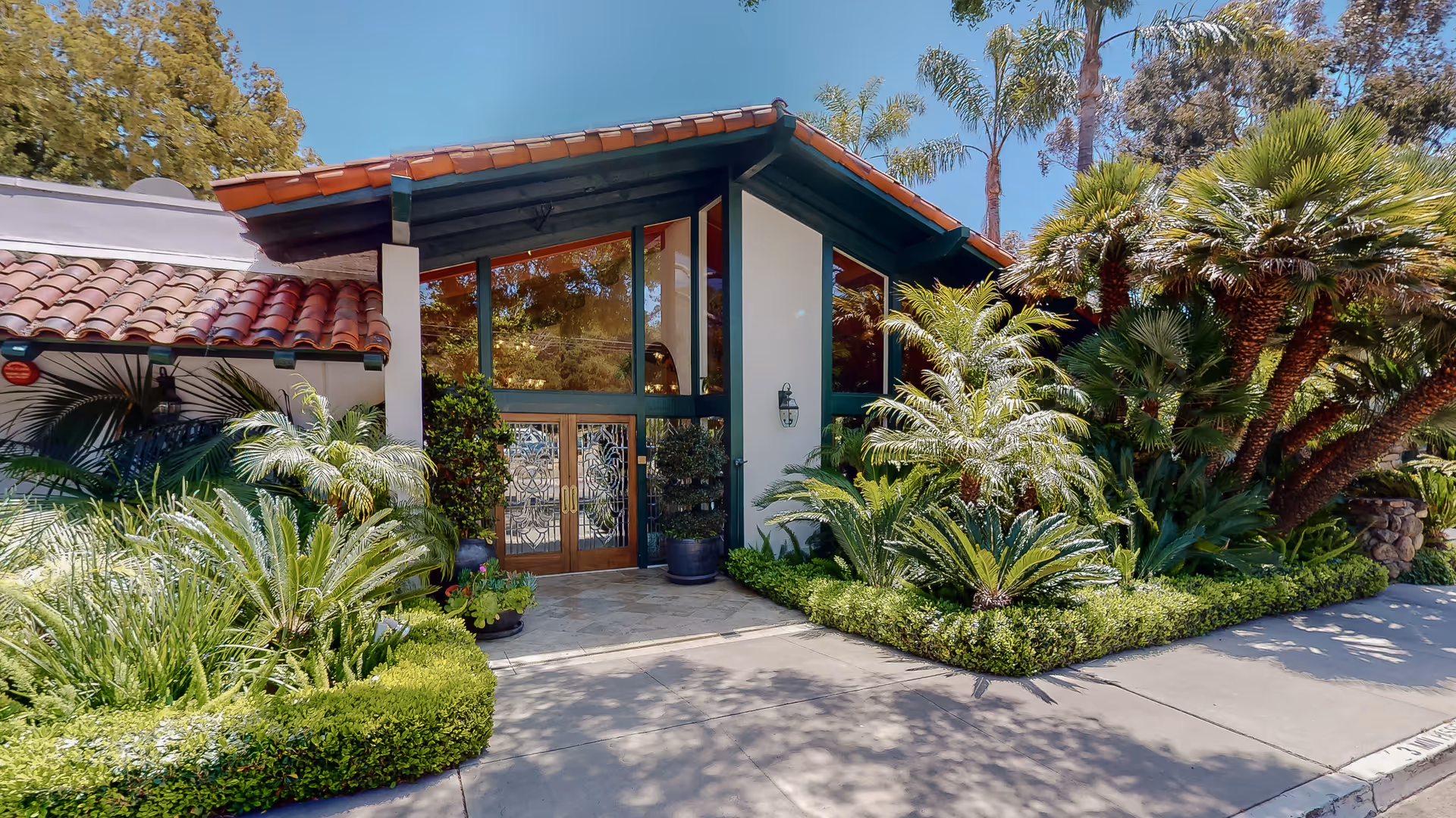 Entrance of Mission Park Healthcare Center featuring a building with a sloped roof and large glass windows. The entrance has double wooden doors with decorative glass panels, surrounded by lush green plants and palm trees. The area is well-lit with natural sunlight and has a clean concrete pathway leading to the doors.